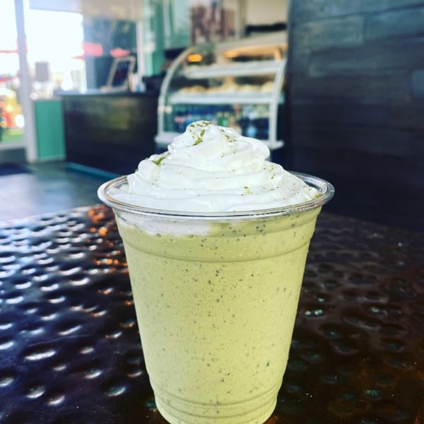 Green smoothie with whipped cream and green powder on a dark textured table inside a restaurant.