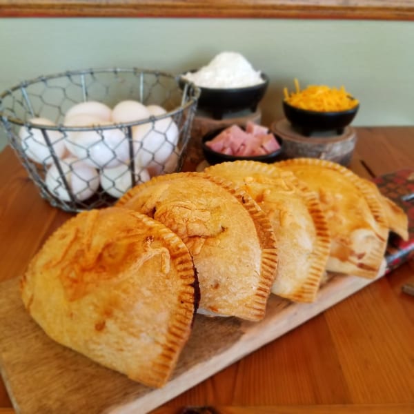 Golden-brown empanadas on a wooden board with ingredients like eggs, flour, ham, and cheese.