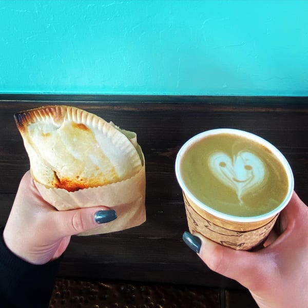 Hands holding a Best Artisan Empanada in a paper sleeve and a latte with heart art.