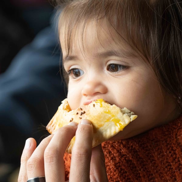 A child with dark hair eats an empanada, held by an adult hand.