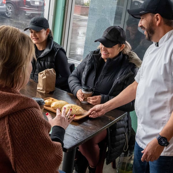 A chef serves Best Artisan Empanadas to three smiling women at a rustic wooden table.